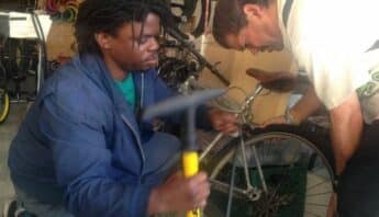 A black man kneeling next to a bike and holding a bike pump with another man bent over the bike