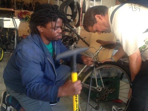 A black man kneeling next to a bike and holding a bike pump with another man bent over the bike
