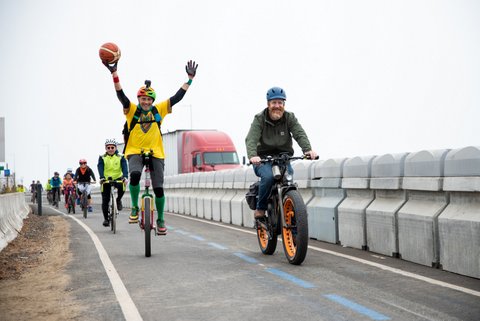 Cyclists on a bike lane separated from vehicular traffic by physical barriers. One cyclist is riding with his arms in the air. A semi truck is partly visible on the other side of the barrier.