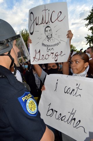 Protesters with signs that say "Justice for George" and "I Can't Breathe" face cop in riot gear.