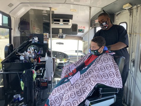One man cuts another man's hair, both wearing medical masks, in barber chair on a bus