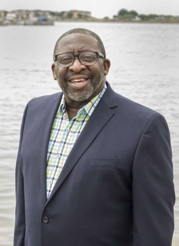 A Black man in a suit with no tie and glasses standing in front of a body of water.