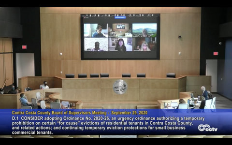 Board of Supervisors meeting with some distanced in government building and others shown on big screen.