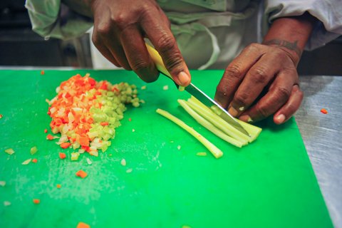 Close up of a Black person's hands cutting thin strips of celery next to a pile of minced carrots and celery.