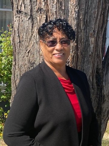 A Black woman wearing tinted glasses, red shirt and black jacket standing in front of a tree.