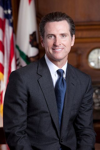 Gavin Newsom, a white man in dark suit with blue tie, with U.S. and California flags over his right shoulder.
