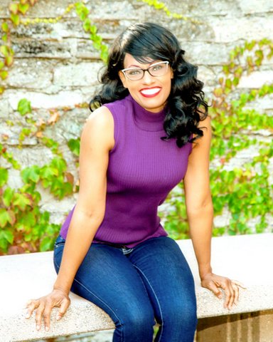 Janet Dandie, a smiling Black woman wearing a purple sleeveless top, blue jeans and glasses.
