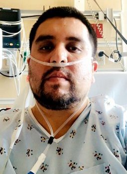 A Latino man in a hospital bed and gown with oxygen tube in his nose.