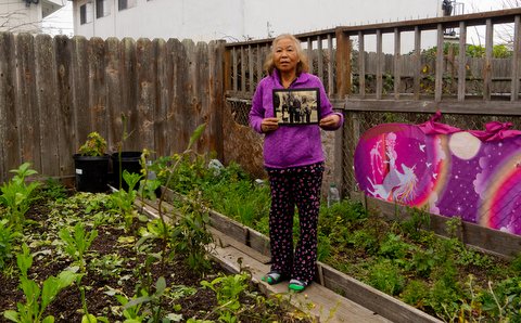 An older Asian woman holding an old photo stands in a garden next to a pink screen with rainbow and unicorn designs.