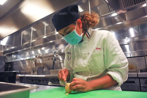 A woman with curly hair, black cap, blue mask and white chef's coat cuts a potato.