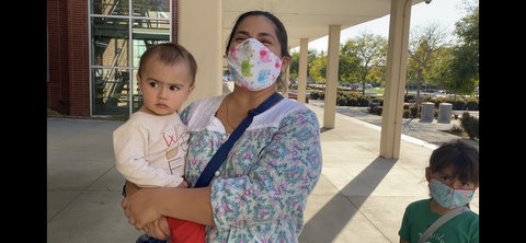 A woman wearing a face mask and holding a baby with a small child wearing a mask next to her.