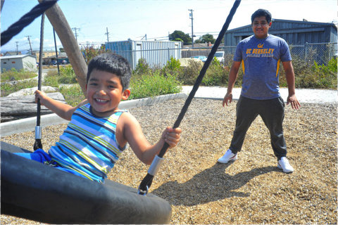 A smiling little boy on a tire-shaped swing in a park with his dad, wearing a UC Berkeley T-shirt, standing a few feet behind him.