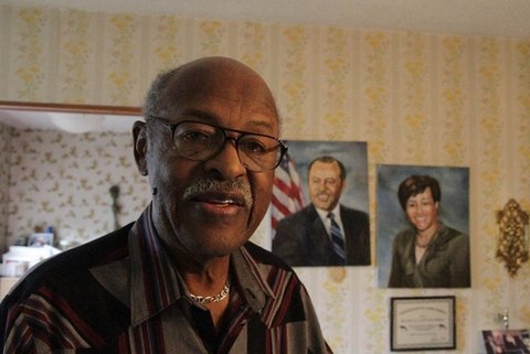 An older balding Black man wearing glasses with paintings of a Black man and woman behind him.