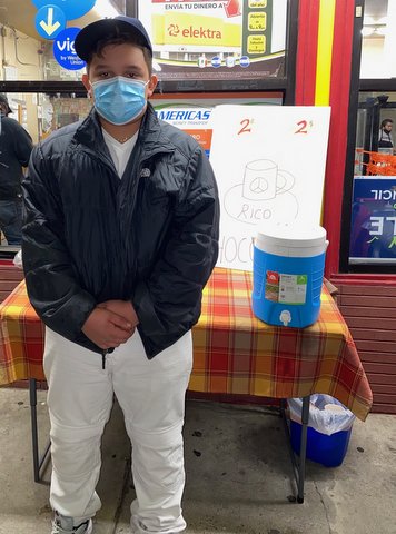 Boy in coat, face mask and cap stands in front of a small table in front of a business.