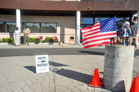 A U.S. flag, a sign that says "Vote" and "Votar" with an arrow and people in masks outside a building.