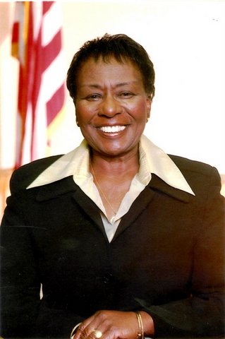 Irma Anderson, a smiling Black woman in a suit, with U.S. flag in background