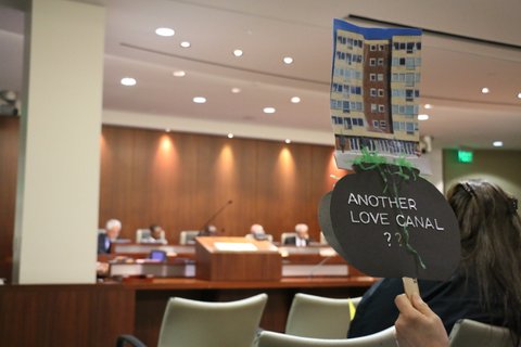 Person at City Council meeting holding up picture of building with words "Another love canal?"