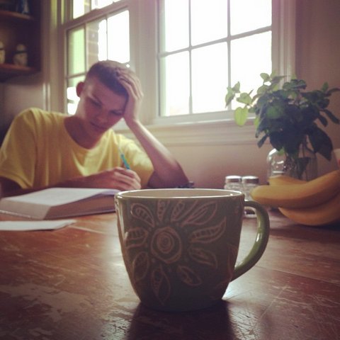 Young man in yellow T-shirt at a table, looking at a textbook and holding his head in one hand.