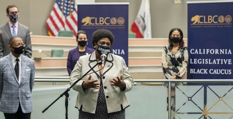 Black woman wearing at a microphone wearing mask that says "California Safe." Behind her are four people in masks spread out and a California Black Legislative Caucus sign.