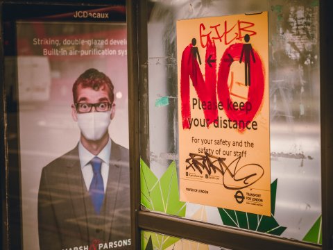 "NO" in big red letters and graffiti on sign that says "please keep your distance" next to poster with man in face mask