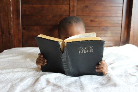 An African American boy in bed reading the Bible.