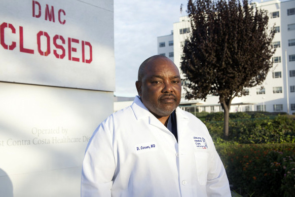 Black man in lab coat near wall that says "DMC CLOSED"
