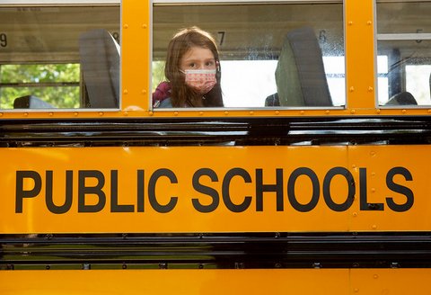 Girl in face mask looks out the window of bus that says "public schools"