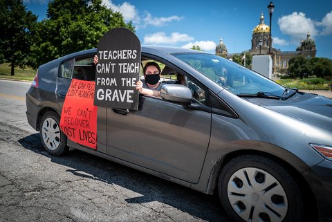 Masked woman holding tombstone-shaped sign out car window that says "Teachers can't teach from the grave."