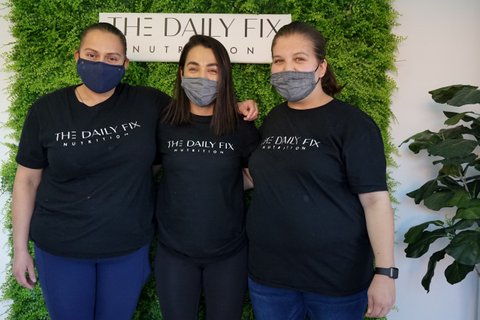 Three Latinas in "The Daily Fix Nutrition" T-shirts in front of a plant wall with a Daily Fix sign.