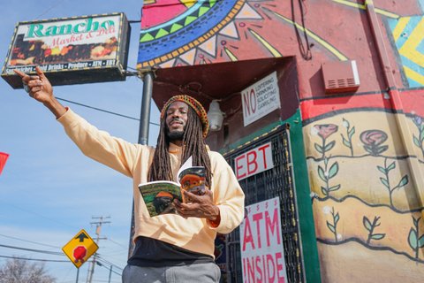 Donté Clark in front of Rancho Market, holding book with other arm outstretched.