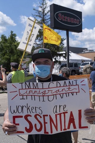 Man in blue surgical mask holds sign that says "Immigrant workers are essential."