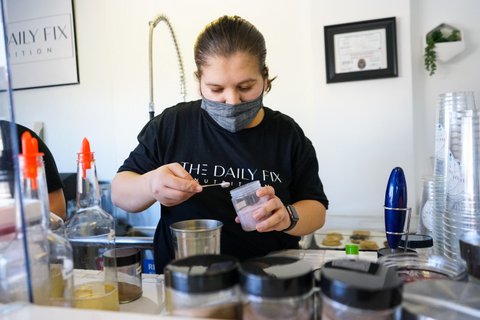 Woman in face mask and Daily Fix Nutrition T-shirt with hair pulled back scoops powder while preparing a drink.