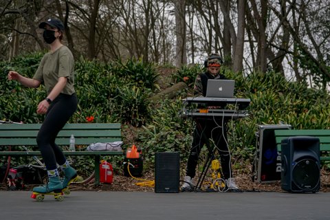 DJ between park benches with trees in background and skater in a mask in the foreground.
