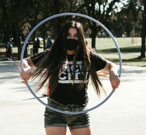 Long-haired woman in mask holding a hula hoop vertically in front of her.