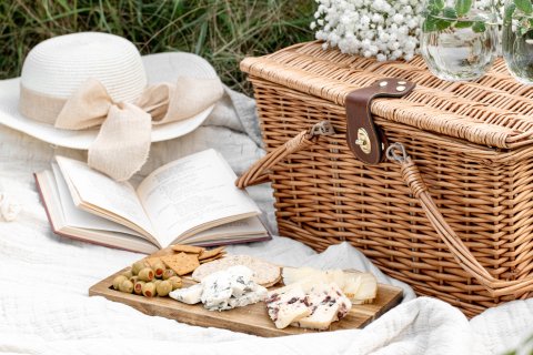 Picnic basket, hat, open book and cheeseboard on a blanket.