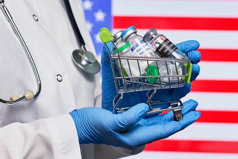 Closeup of vials in tiny shopping cart held by person with lab coat, stethoscope and blue-gloved hands against U.S. flag background