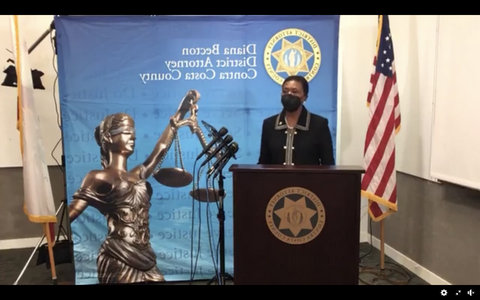 Black woman at lectern with U.S. flag and picture of Lady Justice behind her.