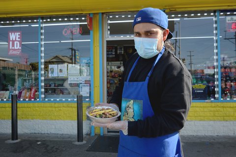 Man wearing face mask, gloves and blue apron and backwards baseball cap holds a dish of food outside a market.
