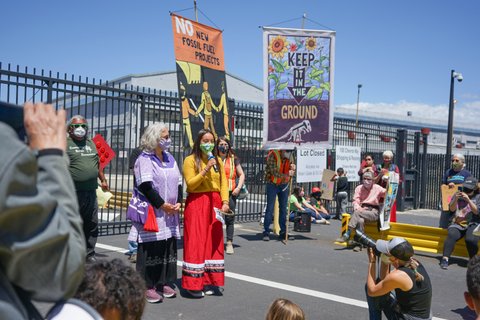 A young woman in a mask, yellow top and red skirt speaks into a microphone among a crowd of people. Signs nearby read "no new fossil fuel projects" and "keep it in the ground."