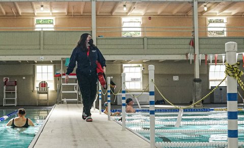 A female lifeguard wearing a mask and carrying a board looks out from a walkway between two pools.