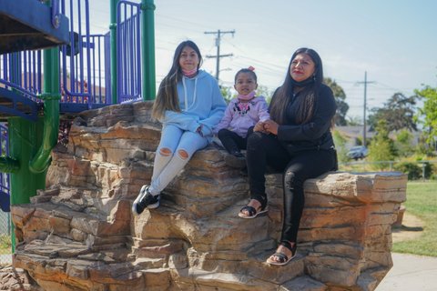A 12-year-old girl, a 4-year-old girl and their mother sitting left to right on rocks in a park.