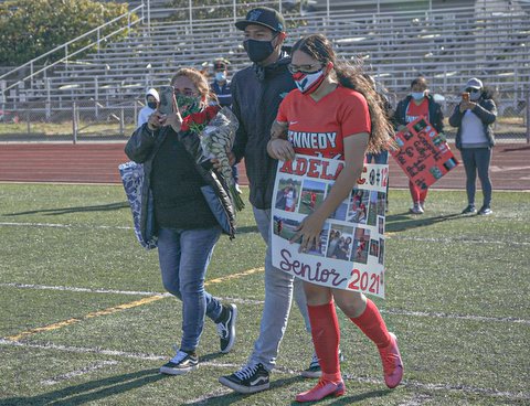 Girl in red soccer uniform and white mask holding sign and walking on field with two people