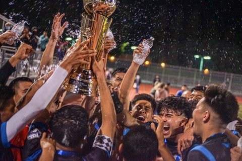 A group of young Latino men celebrate with arms, water bottles and a trophy raised. A spray of water is visible above them.