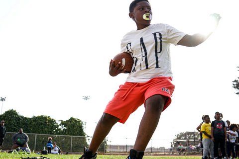 A young Black boy wearing a white T-shirt with "GAP" in big letters and orange shorts with a football under one arm and the other outstretched