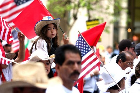 A Latina little girl on an unseen person's shoulders among a crowd of people holding U.S. and red flags