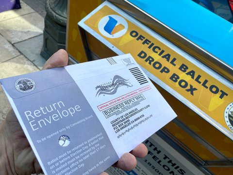 Hand holding a sealed envelope marked "official election mail" in front of an official ballot drop box