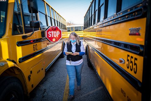 An older white woman in medical mask walks between two school buses, one of which has its stop sign out