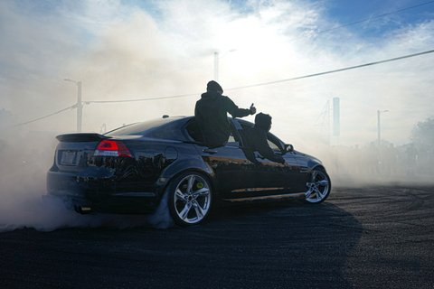Black car in a smoky setting with one passenger sitting on a doorframe and another leaning out a window