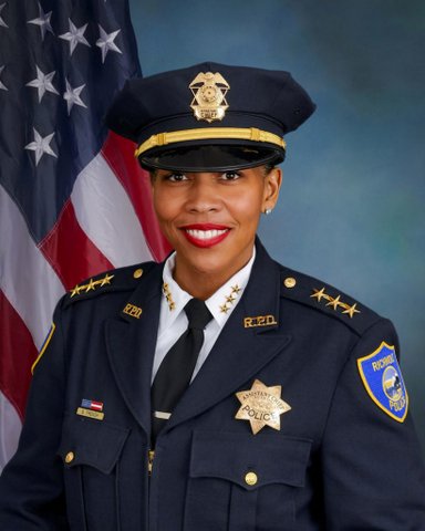 A Black woman police officer in dress uniform with badge that says "assistant chief" in front of the U.S. flag.
