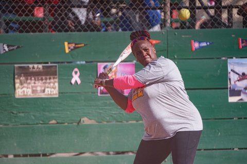 A Black woman at bat during a softball game. She is wearing a gray T-shirt over a top with pink long sleeves.